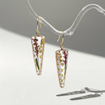 Gold hook earrings with clear resin pendants encasing dried flowers and leaves, displayed under natural sunlight.