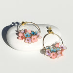 “Gold hoop earrings with pink, blue, and lilac enamel flowers, handcrafted floral design displayed on white stone.