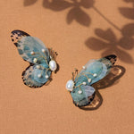 Green butterfly statement earrings with delicate translucent wings and freshwater pearl details, placed on a textured tan background