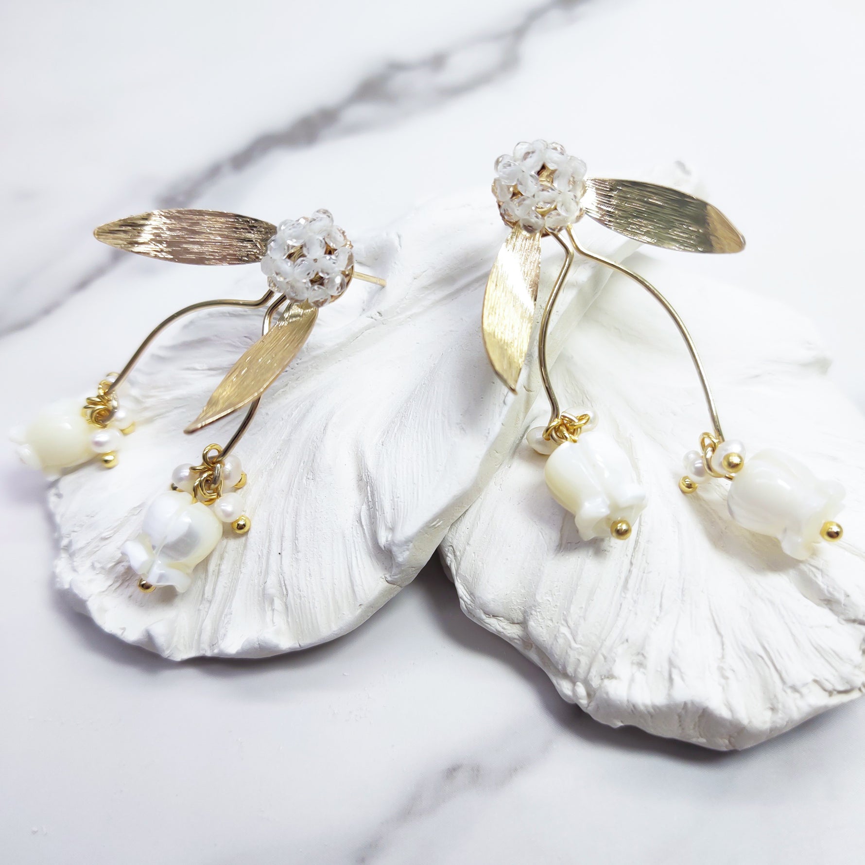 A pair of gold-plated stud earrings featuring hand-carved mother-of-pearl tulip drops, delicate freshwater pearls, and beaded glass floral studs with gold leaf accents, displayed on white textured petals.