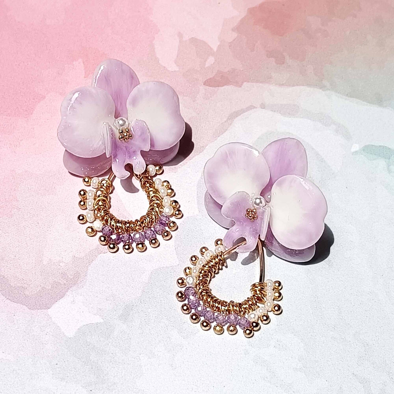 Pair of light purple orchid earrings with gold wire hoops, adorned with tiny pearls and lavender beads, displayed on a soft pink watercolor background.