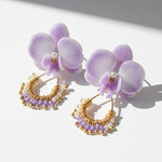 Gold hoop earrings with purple accents next to a purple orchid on a white background