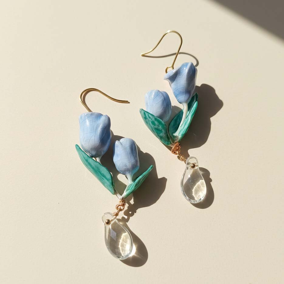 Blue tulip-shaped earrings with green leaves and clear teardrop pendants on a beige background