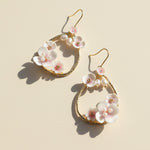 Gold teardrop earrings adorned with delicate pink and white enamel flowers and small pearls, photographed in soft sunlight.