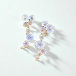 Close-up of white and lilac floral pearl drop earrings on soft white surface, gold-plated detailing.