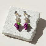 Hand-painted enamel grape and pearl drop earrings on white stone background, sunlight and shadow.