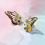Gold butterfly earrings with green gemstones, pearls, and floral accents on pastel pink background.