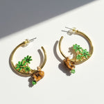 Gold hoop earrings with green leaf and brown sloth charms on a white background