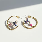 Gold hoop earrings featuring hand-painted hummingbirds sipping from pink flowers, captured in natural sunlight.