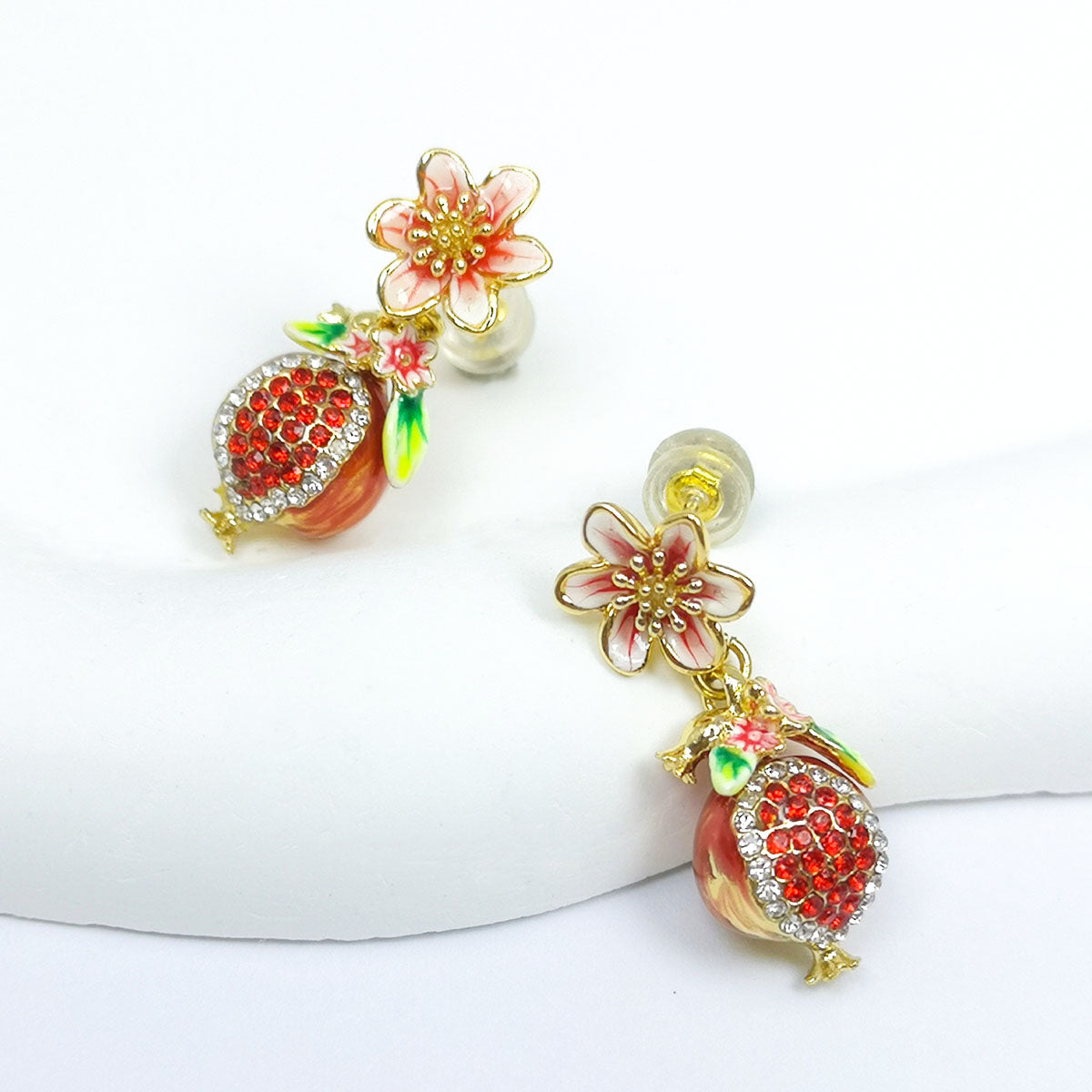 Close-up of hand-painted enamel pomegranate and flower stud earrings with red cubic zirconia and gold-plated brass, displayed on a white surface.