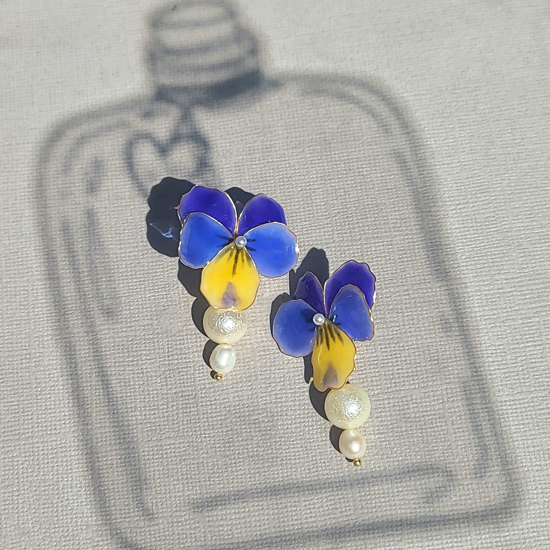 A pair of enamel earrings featuring vibrant blue and yellow pansy flowers, accented by a small white pearl at the center. Two textured and smooth pearls dangle from each floral motif, creating a graceful, feminine look. The earrings are photographed outdoors on a textured grey fabric background, with soft shadows of a bottle-shaped object framing the image and creating a charming, artistic effect.