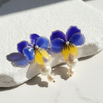 Hand-painted blue and yellow pansy flower earrings with pearl accents, displayed on a textured white surface.