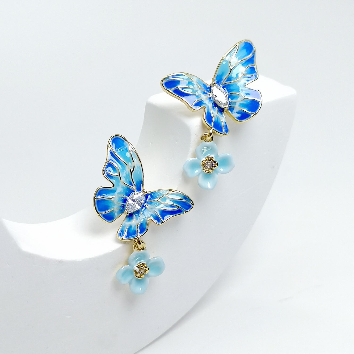 A pair of gold-plated stud earrings featuring hand-painted blue enamel butterflies with sparkling cubic zirconia centers and dangling pastel blue flower charms.