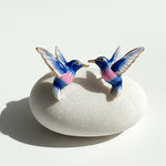 Close-up of blue and pink enamel hummingbird earrings displayed on white stone, natural light.