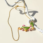 Gold chain necklace featuring a hand-painted tulip pendant with pink, peach, and green enamel flowers and leaves, displayed in sunlight on a cream background