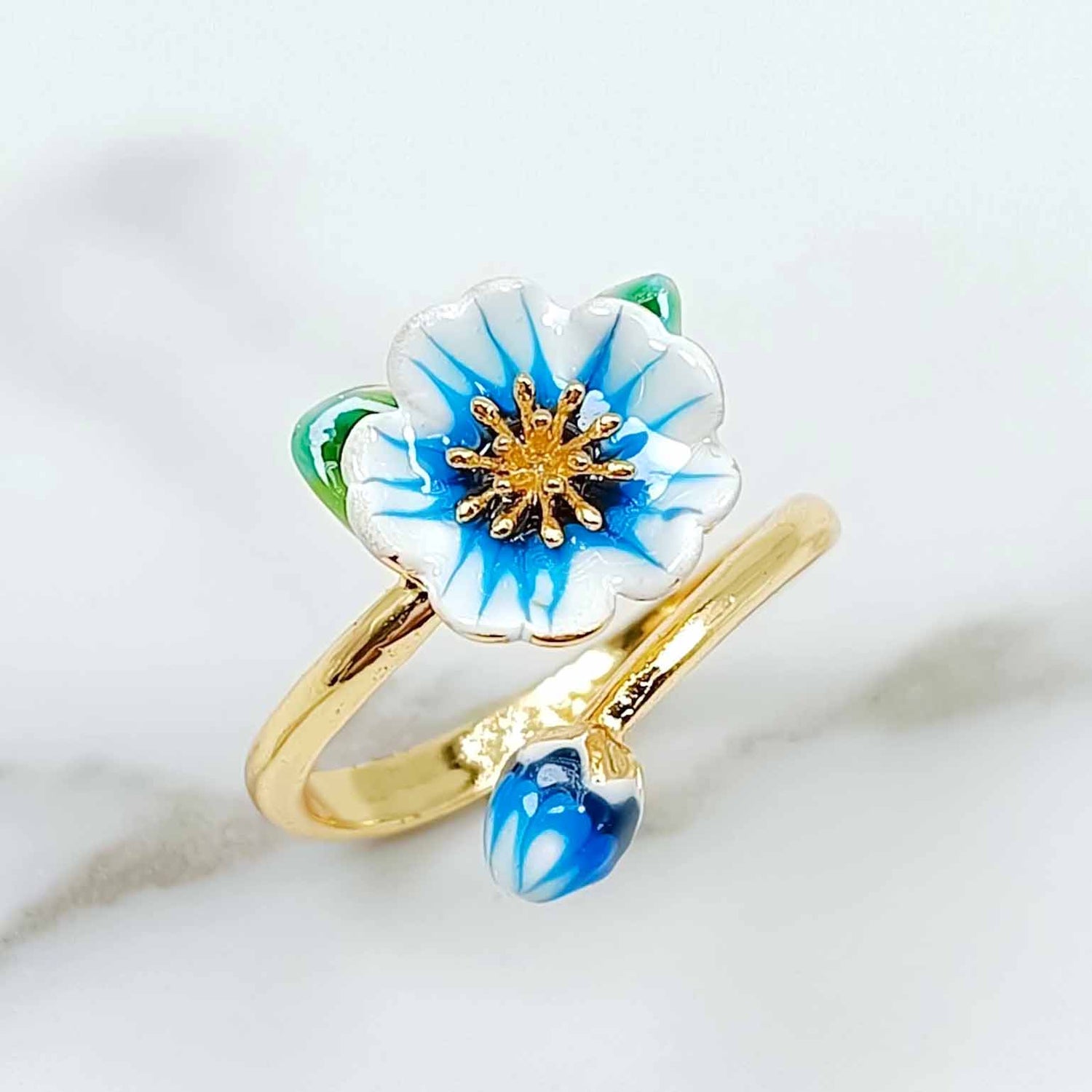 Gold-plated adjustable ring featuring a hand-painted blue and white enamel flower with a matching bud and green leaf detail on white marble background.