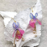 A pair of Iris flower dangle earrings with pink and purple petals and gold-colored chains, featuring fresh water pearls, displayed on a white leaf-like surface.