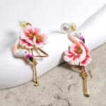 Asymmetrical pink flamingo enamel earrings with gold-finished legs, pink-white flower detail, and pearl stud backings, displayed on marble and ceramic props.