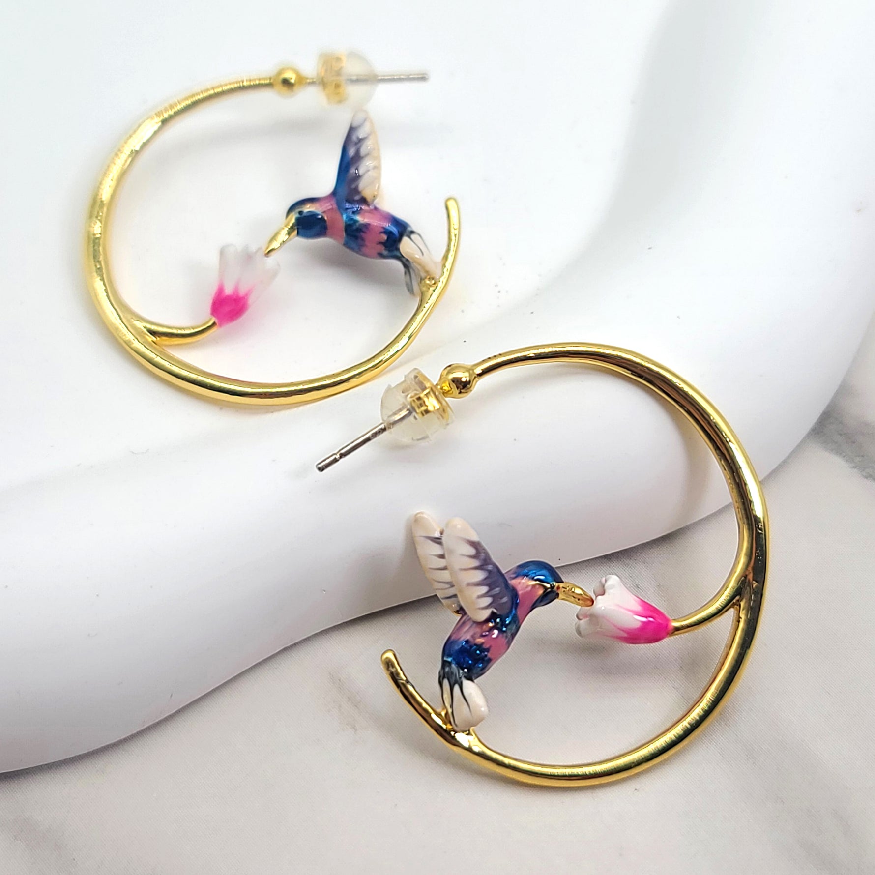 A pair of charming hoop earrings featuring enamel-painted hummingbirds feeding on pink and white flowers. Crafted from brass and 18k gold-plated for a luxurious finish, these earrings have a diameter of 3.2cm (1.25”) and include silver posts for comfortable wear. The design captures the dynamic elegance of hummingbirds in flight, offering a whimsical and colorful statement accessory.