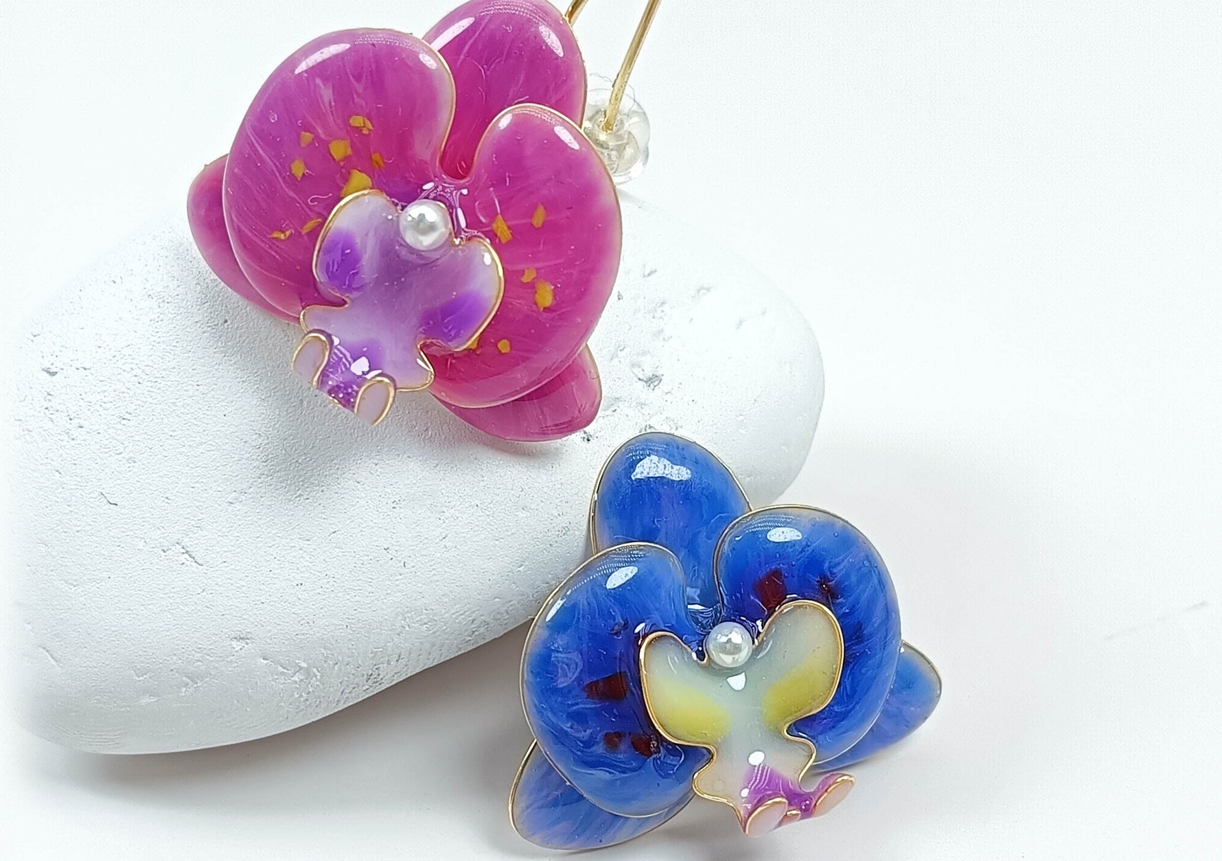 Hand-painted orchid flower earrings in fuchsia pink and electric blue with pearl centers, displayed on a white stone background