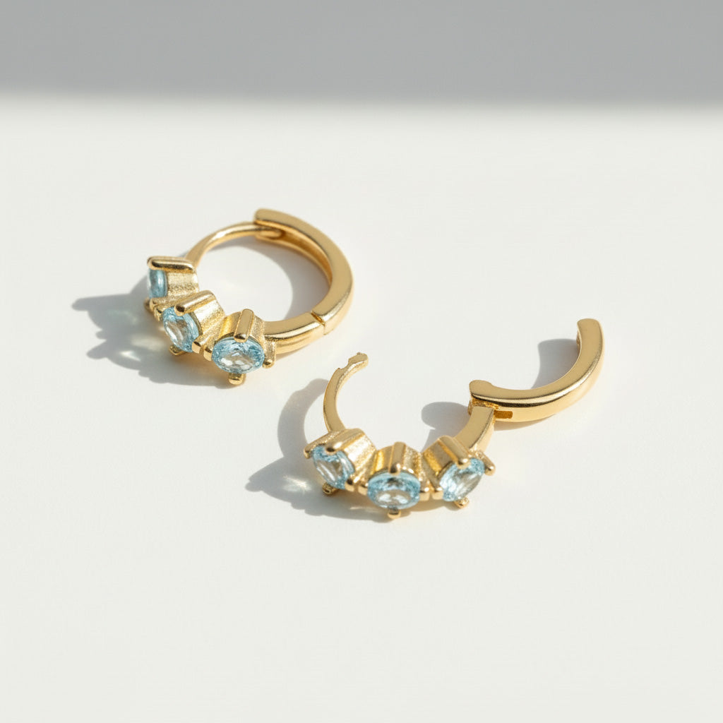 Gold-plated silver hoop earrings with three light blue cubic zirconia birthstones, elegant minimalist design.