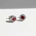 Silver stud earrings with platinum plating and garnet birthstone center, surrounded by clear CZ halo.