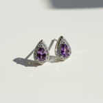 Teardrop stud earrings with purple birthstone, platinum-plated silver setting, and halo of clear cubic zirconia.