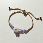Gold adjustable bracelet with amethyst beads and carved white flower charm featuring a purple gemstone centre.