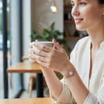Model wearing gold adjustable bracelet with purple beads and white flower charm while holding a cup of coffee.