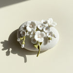White porcelain flower brooch with hand-painted enamel leaves and pearl details, styled on stone surface