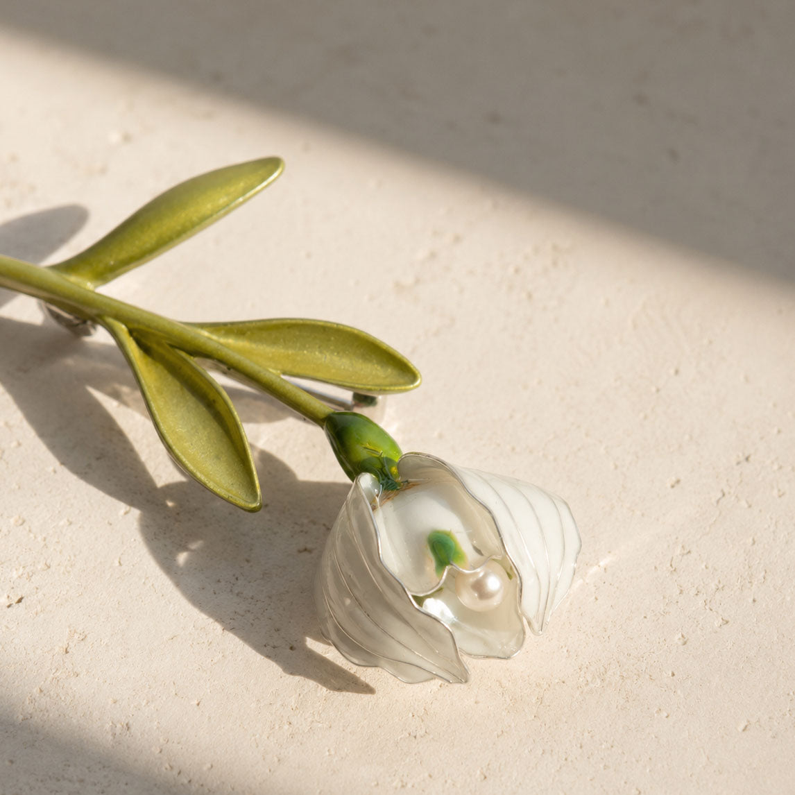 Lily of the valley pearl brooch with hand-painted enamel flower resting on a light stone surface.