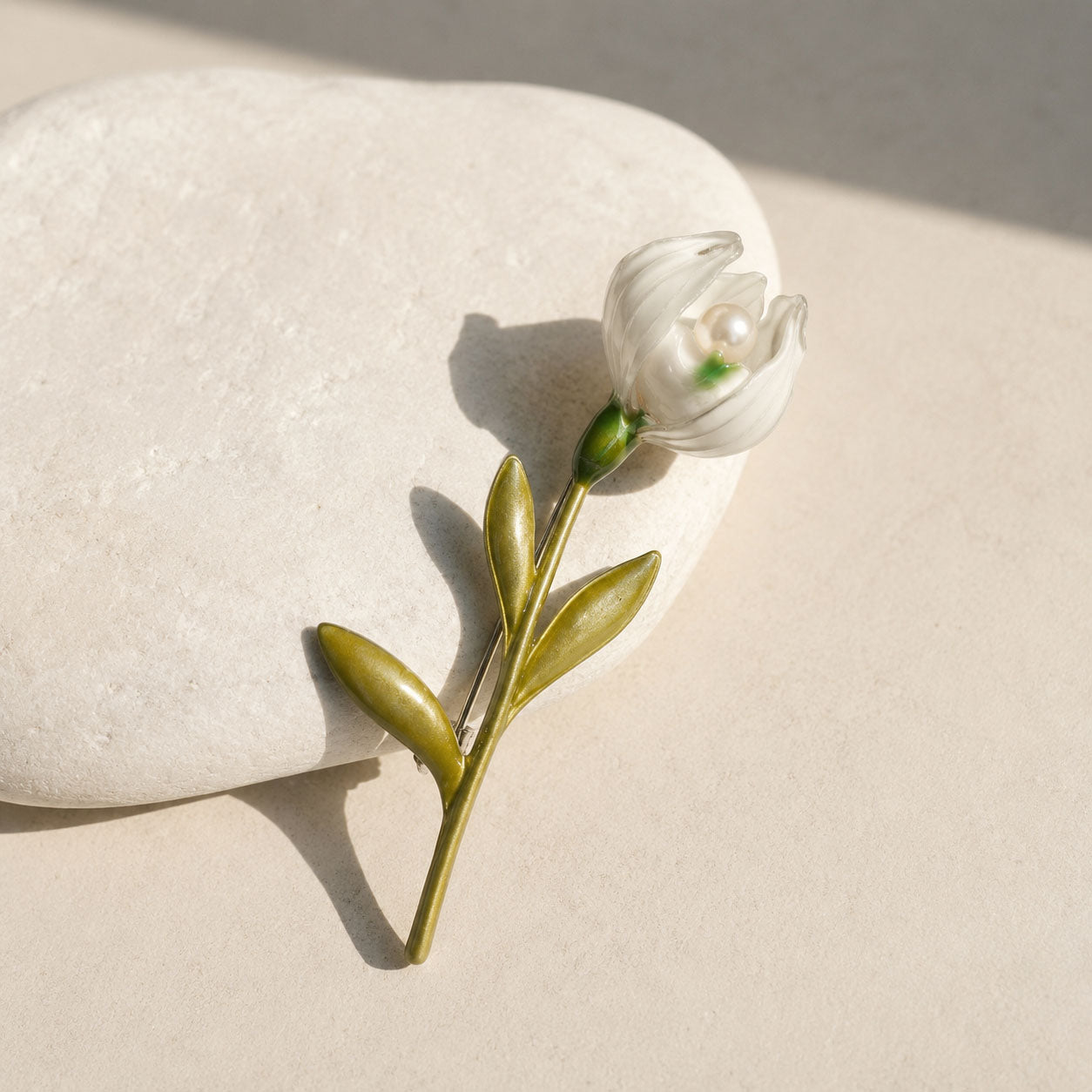 Handcrafted lily of the valley pearl brooch with white enamel bloom and green leaves on a smooth white stone.
