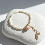 Freshwater pearl bracelet with hand-painted enamel peach charm in gold-plated brass on a white stone surface