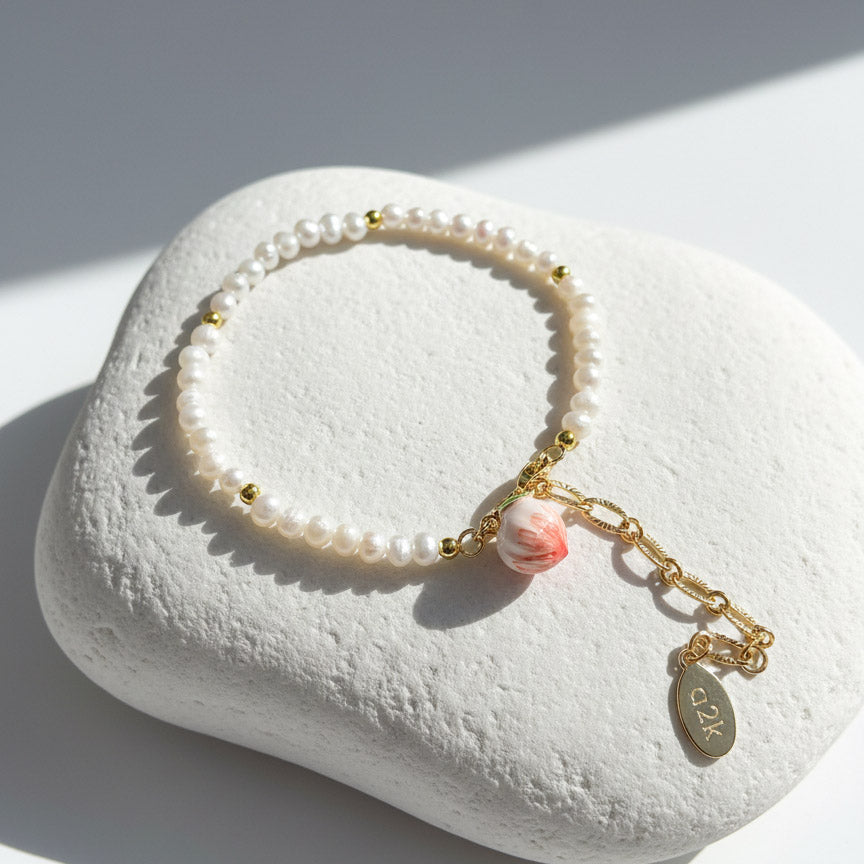 Freshwater pearl bracelet with hand-painted enamel peach charm in gold-plated brass on a white stone surface