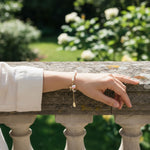 Hand wearing a freshwater pearl bracelet with hand-painted enamel peach charm on a stone railing with a garden background
