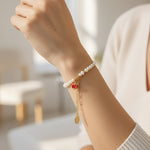 Hand wearing a freshwater pearl bracelet with pavé red pomegranate charm against a blurred indoor background