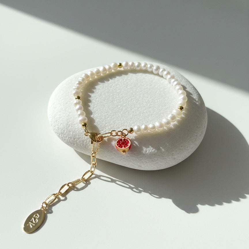 Freshwater pearl bracelet with pavé red pomegranate charm in 18K gold-plated brass on a white stone surface