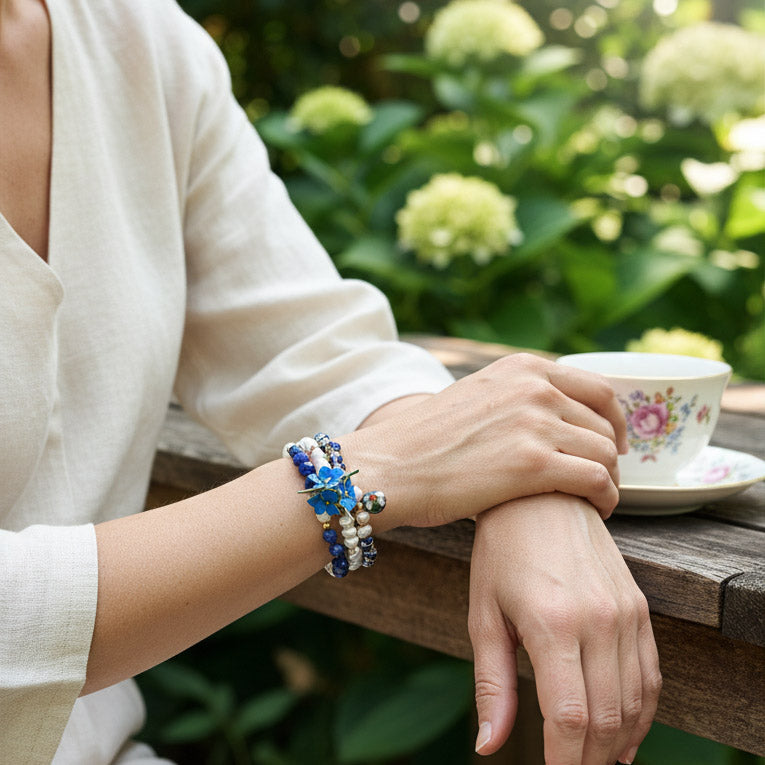 Person holding a teacup with floral design, wearing colorful Layered sodalite lapis and cloisonné beaded bracelet set on a wooden table with greenery in the background.