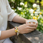 Person holding a cup of tea with a yellow opal tiger eye and freshwater pearl floral beaded bracelet set with greenery in the background