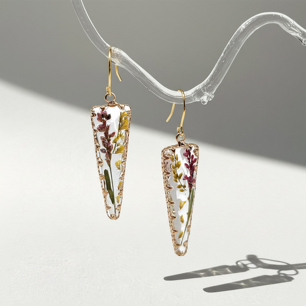 Gold hook earrings with clear resin pendants encasing dried flowers and leaves, displayed under natural sunlight.