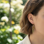 Close-up of a person wearing a gold earring with green gemstone against a blurred natural background
