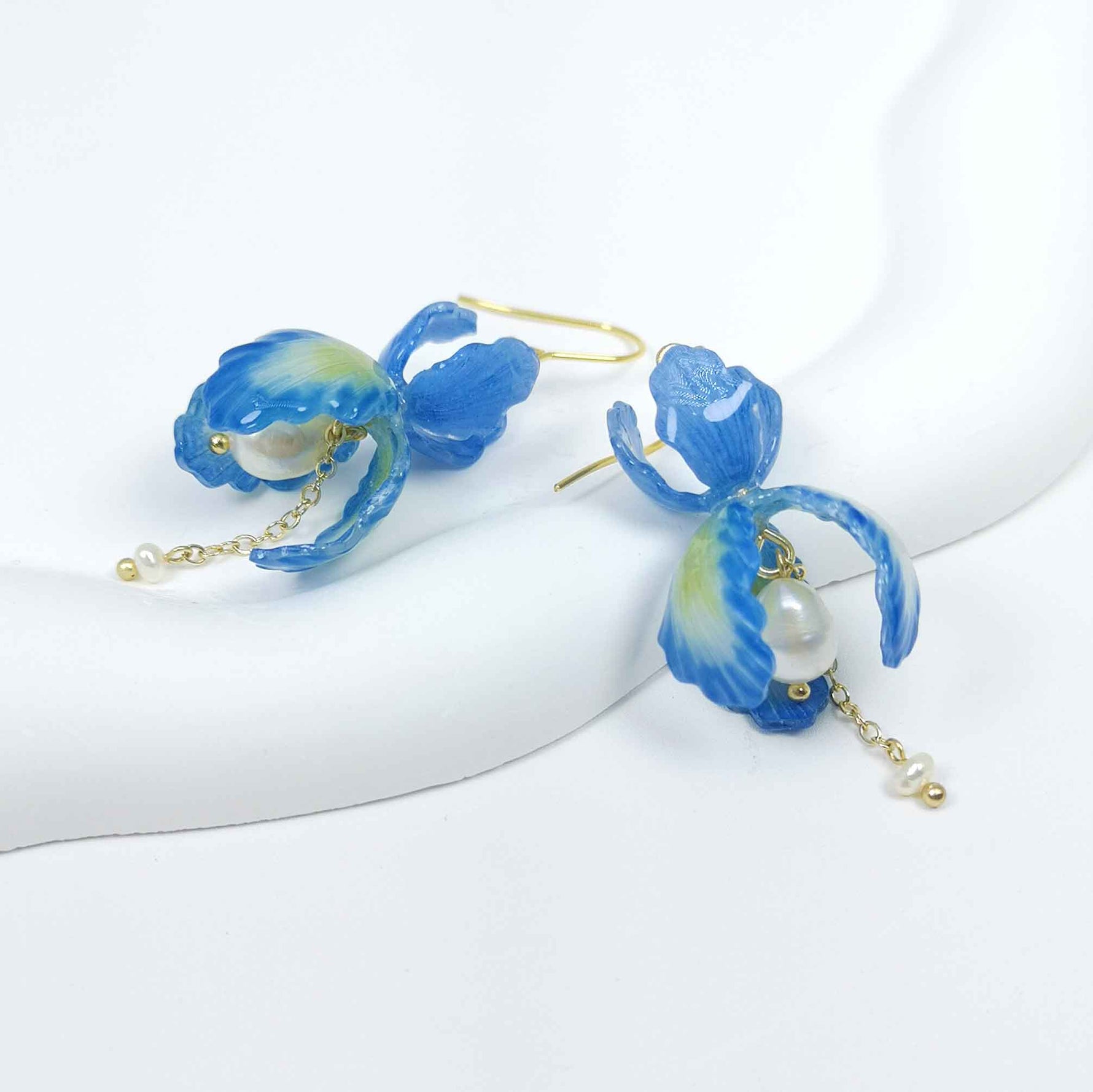 Blue iris flower drop earrings with handpainted resin petals, gold-plated hooks, faux pearl center, and dangling freshwater pearl chain on white background