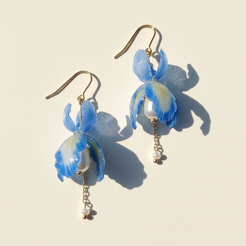Blue iris flower drop earrings with pearls and gold chains, handcrafted resin floral jewelry on light background.