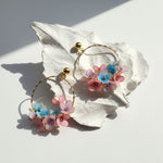 Gold hoop earrings with hand-painted blue and pink enamel flowers, displayed on white leaf background in sunlight.