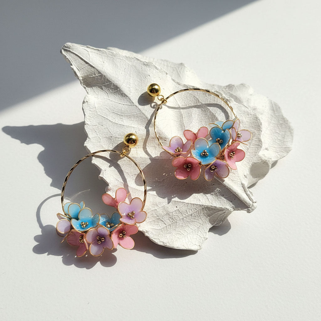 Gold hoop earrings with hand-painted blue and pink enamel flowers, displayed on white leaf background in sunlight.