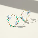 Gold hoop earrings with blue butterfly and green leaf designs on a light background