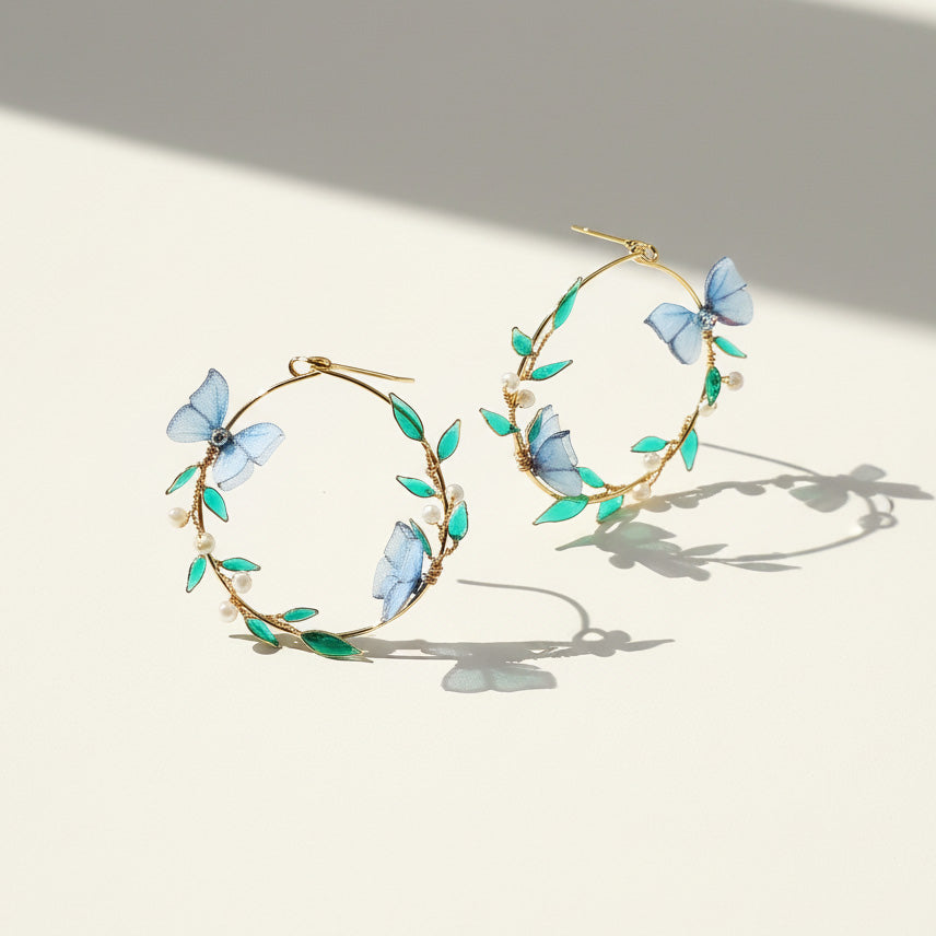 Gold hoop earrings with blue butterfly and green leaf designs on a light background