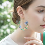 Close-up of a woman wearing gold hoop earrings with blue butterfly and green leaf designs.