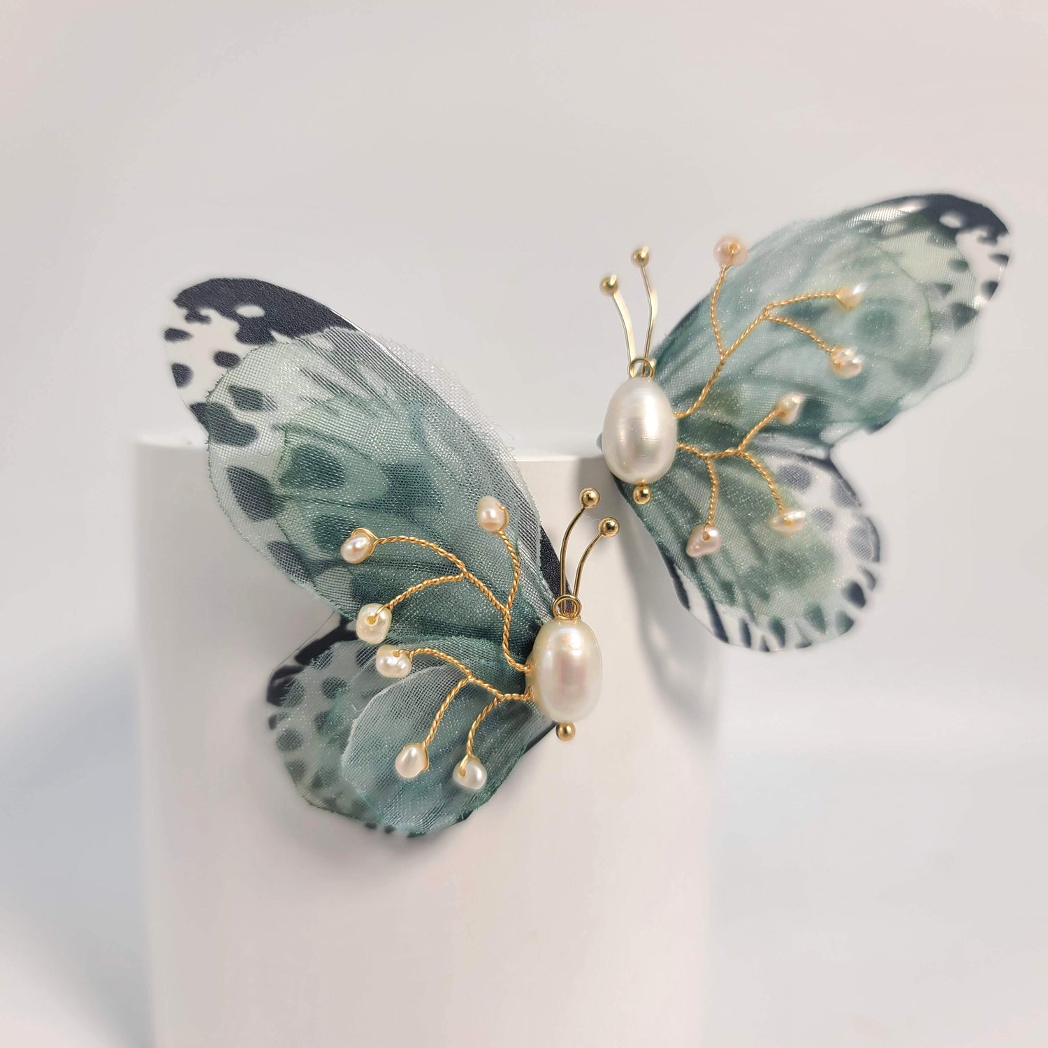 Green butterfly earrings with intricate gold wire details and scattered pearls, styled on a neutral beige textured backdrop.