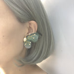 Close-up side profile of a woman wearing a large butterfly earring with green wings, freshwater pearls, and golden accents. The woman's hair is styled in a sleek, gray bob.